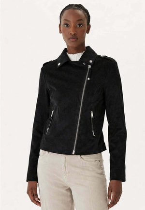 Donna che indossa una giacca asimmetrica in camoscio nero con cerniera sopra una maglietta bianca e pantaloni beige chiaro, in piedi davanti a uno sfondo semplice.