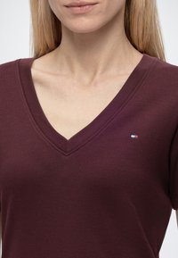 Γυναίκα φοράει σκούρο μαρόν V-neck πουκάμισο με μικρό ορθογώνιο κόκκινο, άσπρο και μπλε λογότυπο στο αριστερό στήθος.