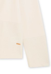 Top de manga larga color beige claro con panel inferior texturizado, que presenta una sección superior suave y un pequeño detalle de logo metálico.