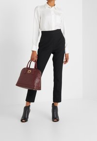 Burgundowa pikowana torebka z złotymi akcentami, okrągły kształt i podwójne uchwyty. Modelka ma na sobie białą koszulę i czarne dopasowane spodnie oraz czarne botki na obcasie.