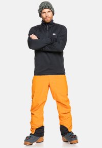 Mustat fleece-takkipuserot, oranssit hiihtohousut ja harmaamustat hiihtokengät. Fleecessä on teksturoitu pinta ja housuissa on rento leikkaus.