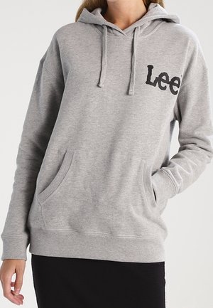 Mujer con sudadera con capucha gris claro de Lee con bolsillo frontal y cordones, con la mano en el bolsillo, combinada con una falda negra.