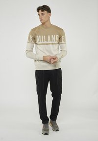 Maglione a righe beige e crema con testo "MILANO", abbinato a pantaloni neri e sneaker grigie con dettagli multicolori.