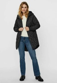 Cappotto lungo imbottito nero con cappuccio, dotato di zip frontale, tasche laterali e orlo curvo, abbinato a un maglione crema e jeans blu.