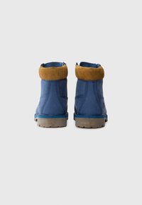Une paire de bottes de cheville en cuir bleu avec des cols rembourrés marron et des semelles en caoutchouc beige, présentée de dos sur un fond blanc.