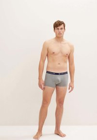 Graue Baumwoll-Boxershorts mit einem marineblauen Bund, auf dem der Markenname "TOM TAILOR" prangt. Das Modell steht barfuß vor einem neutralen Hintergrund.