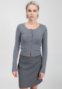 Cardigan grigio lavorato a maglia con maniche lunghe e bottoni frontali, abbinato a una gonna grigia scura con piccole pois bianchi e dettagli in metallo.