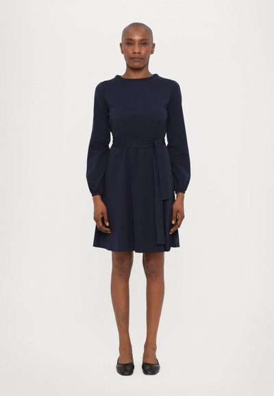 Robe bleu marine avec des manches longues, un col rond et une ceinture assortie à la taille. La jupe plissée ajoute du volume. Texture de tissu lisse.