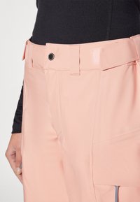 Light pink outdoor pants with a button closure, belt loops, and side pockets featuring zippers, made of a durable, textured fabric.