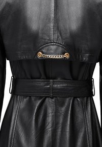 Zwarte leren trenchcoat met een ceintuur in de taille. Opvallende details zijn onder meer een gouden kettingaccent op de rug, een soepele textuur en een op maat gemaakte ontwerp.