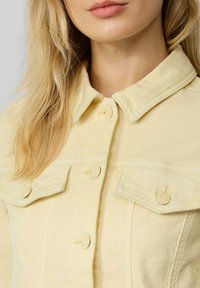 Lichtgele denimjas met een puntige kraag, knoopsluitingen en twee borstzakken met gestikte details. Zachte textuur.