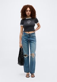 Νεαρή γυναίκα με σγουρά μαλλιά, φορώντας μαύρο κολλητό τοπ με σχέδιο ήλιου, σκισμένο μπλε τζιν, μαύρα σανδάλια, κρατώντας μαύρη τσάντα, στέκεται μπροστά σε λευκό φόντο.