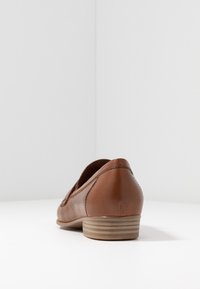 Mocassin en cuir marron avec un petit talon empilé sur une surface blanche et un fond blanc, vu depuis le côté arrière droit.