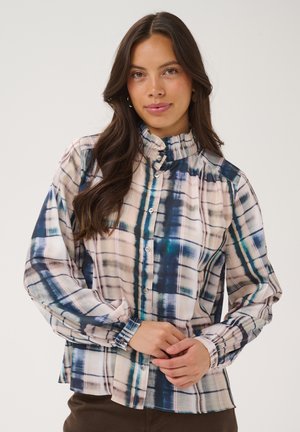 Femme aux longs cheveux bruns portant un chemisier à col montant boutonné avec un motif à carreaux bleu, blanc et beige, debout devant un fond uni.