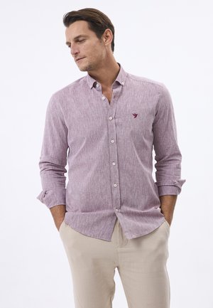 Mężczyzna w jasnofioletowej koszuli z długim rękawem podwiniętym na ramionach i beżowych spodniach, z rękami w kieszeniach, patrzący w lewo.