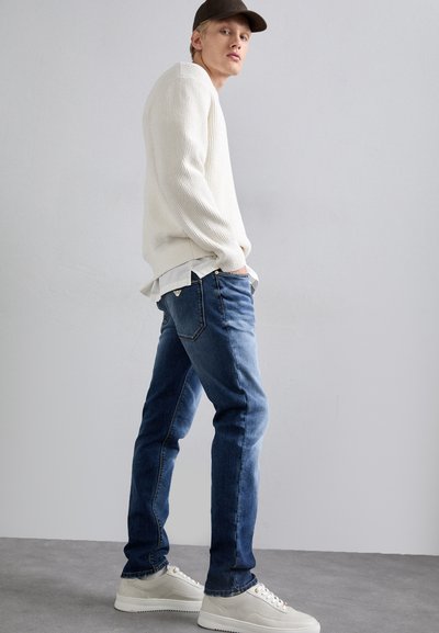 Pulover alb cu dungi, blugi slim-fit în nuanță închisă și adidași deschiși. Modelul se întoarce pe lateral, evidențiind potrivirea și contrastul de culoare al ținutei.