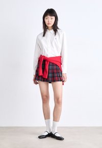Camisa blanca de botones, minifalda a cuadros en rojo, verde y azul marino, suéter de punto rojo atado a la cintura, calcetines blancos y zapatos Mary Jane negros.