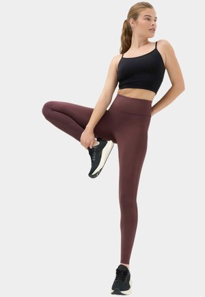 Ung kvinde i sort crop top, mørklilla leggings og sorte sneakers strækker sig ved at holde en fod bag benet mod en ensfarvet baggrund.