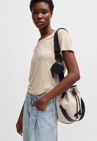 Bucket taske i cremefarvet stof med sorte læderdetaljer, snøre-lukning og justerbar rem. Modellen bærer en beige top og lyseblå jeans.
