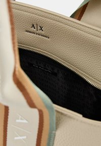 Beige leren tas met textuur, binnenafwerking in zwart, met een ritsvak en geperste branding. Gestreepte stofaccenten zichtbaar.