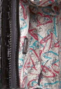 Indvendig i en Guess taske med sort laklæderkant, flerfarvet logo trykt stofforing og en synlig lynlås lomme.