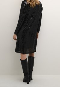 Kvinna i en svart paljettklänning med lång ärm och svarta knähöga läderstövlar med klack, som står mot en ljus, enfärgad bakgrund.