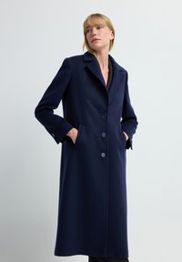 Marineblauer Wollmantel mit Reverskragen, zwei Fronttaschen und Knopfverschluss. Glatte Textur mit einer maßgeschneiderten, langen Silhouette.