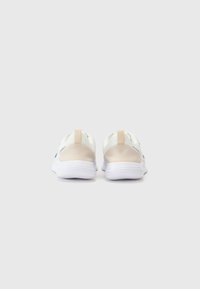 Zapatillas deportivas blancas con un talón de color beige claro, que cuentan con una parte superior de tejido texturizado y una tira de agarre, diseñadas para ofrecer comodidad y soporte.