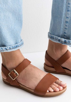 Pies con vaqueros azul claro remangados que llevan sandalias de piel marrón con una hebilla dorada sobre una superficie blanca.