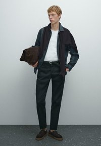 Abbigliamento casual composto da una camicia scura, una maglietta bianca, pantaloni neri e scarpe in suede marroni. Gli accessori includono una borsa marrone e un maglione scuro appoggiato sulle spalle.