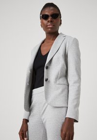 Svijetlosiva sako od teksturirane tkanine s jednim dugmetom, uparen s crnom majicom. Naglašen tamnim dugmadima i uskim krojem.