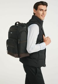 Czarny plecak z wieloma zamykanymi na zamek kieszeniami, teksturowanym materiałem, brązowymi akcentami i usztywnioną konstrukcją, noszony na plecach mężczyzny.