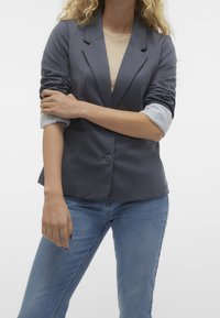 Šedý blazer s klopovými límci a jedním knoflíkem, doplněný o košili s jemným proužkem na rukávech, nošený přes modré džíny.