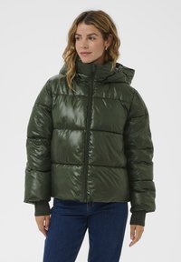 Kvinde iført en skinnende mørkegrøn pufferjakke med hætte, som er lynet op, sammen med blå jeans og strikkede håndledsmanchetter.