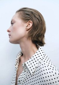 Camicia bianca con pois neri, colletto a bottoni. Tessuto morbido e liscio. Profilo laterale di una persona con capelli corti e ondulati su uno sfondo chiaro.
