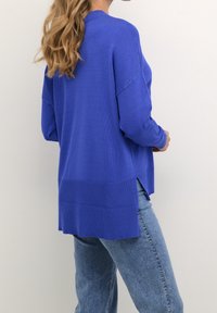 Blauer gestrickter Pullover mit lockerem Schnitt, verfügt über einen gerippten Ausschnitt, lange Ärmel und seitliche Schlitze am Saum. Kombiniert mit hellblauem Denim.