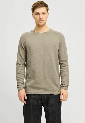 Camicia a maniche lunghe di colore verde oliva in un tessuto morbido e caratterizzato; presenta un collo a giro e maniche raglan, abbinata a jeans in denim scuri.