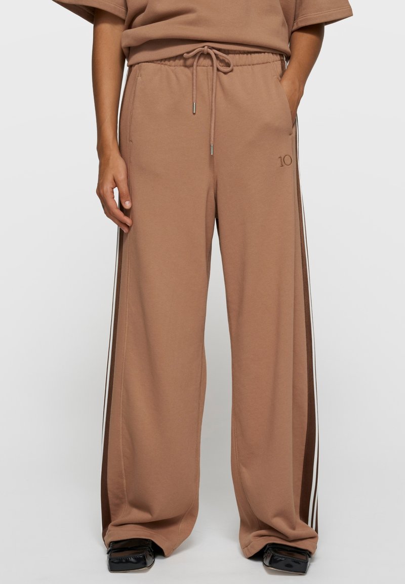 Pantalon de survêtement beige à jambes larges avec une taille élastique, un cordon de serrage, des poches latérales et des rayures verticales contrastées sur les côtés.
