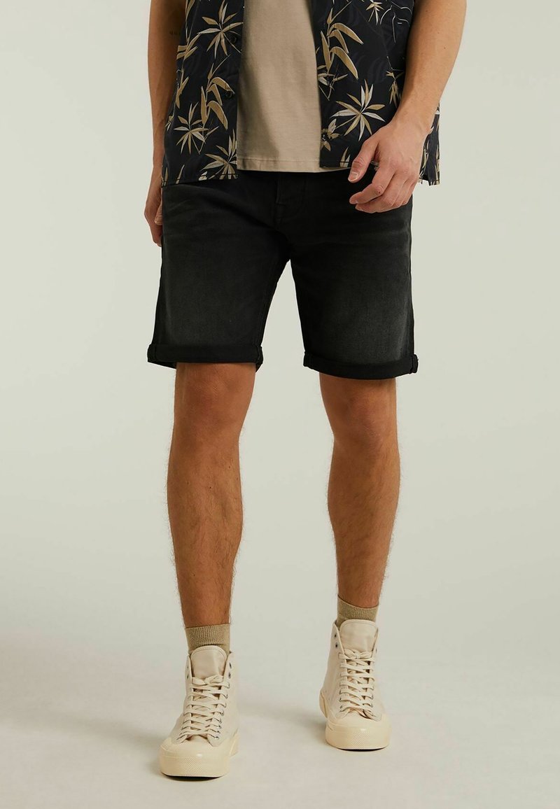 Homme portant un short noir remonté, des chaussettes beige, des baskets montantes crème, un t-shirt beige et une chemise noire à motifs floraux, debout devant un fond uni.