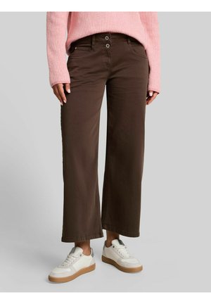 Person trägt braune Hosen mit weitem Bein, einen pinkfarbenen gerippten Pullover und weiße Sneakers mit Gummisohlen, steht vor einem einfarbigen Hintergrund.