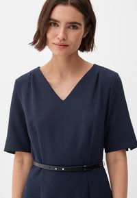 Navyblauw jurk met korte mouwen, V-hals en een zwarte riem. Glad stof met plooien op de schouders en een strakke silhouette.