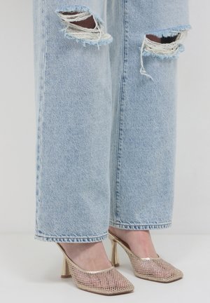 Jean déchiré bleu clair porté avec des mules à talons hauts dorées métalliques texturées sur fond blanc.