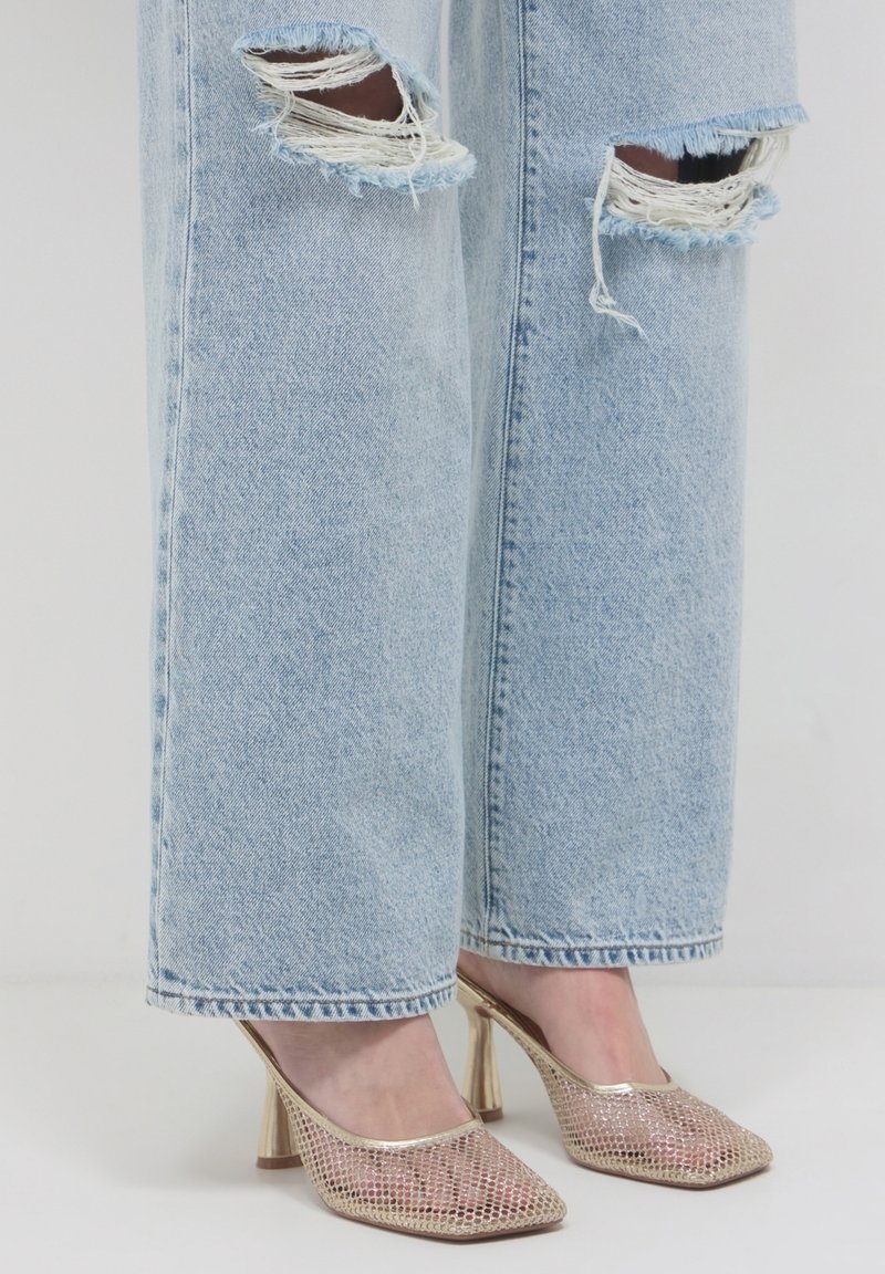 Jean déchiré bleu clair porté avec des mules à talons hauts dorées métalliques texturées sur fond blanc.