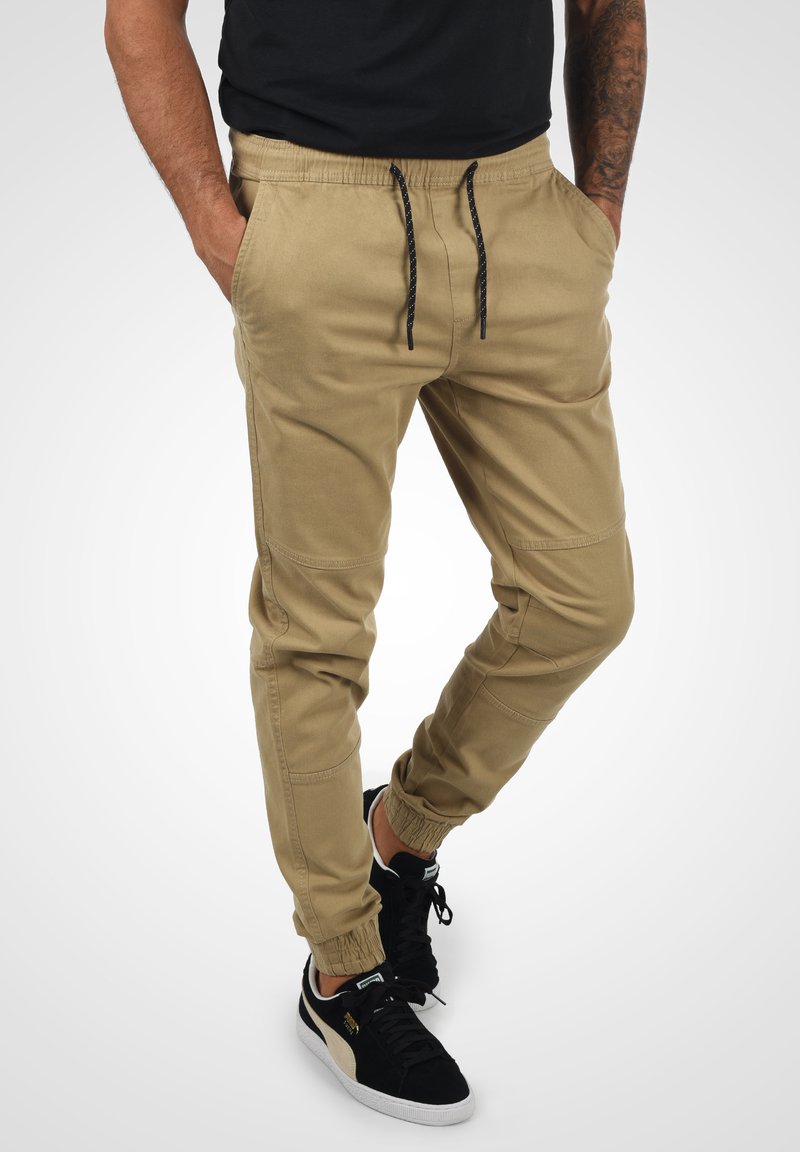 Lys beige joggebukser laget av bomullsblanding, med elastiske mansjetter, sidelommer og en snor i midjen med svarte accenter.
