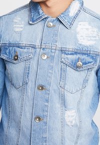 Lichtblauwe spijkerjack met metalen knopen, twee borstzakken en versleten plekken op de kraag, borst en onderkant aan de voorkant.