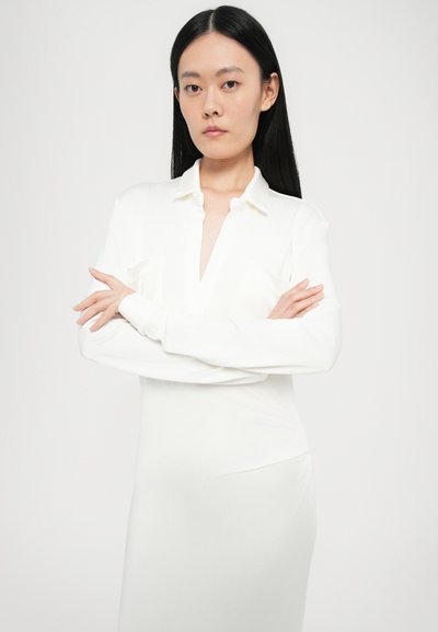 Witte blouse met lange mouwen en een diepe V-hals, knoopsluiting bij de kraag en twee borstzakken. Gemaakt van een gladde, lichte stof.