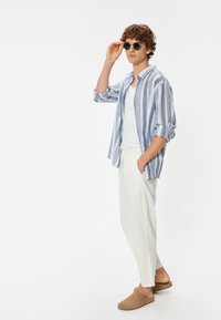 Blauw-wit gestreepte shirt, witte broeken, bruine instapschoenen en ronde zonnebril. Model poseert met één hand op de zonnebril.