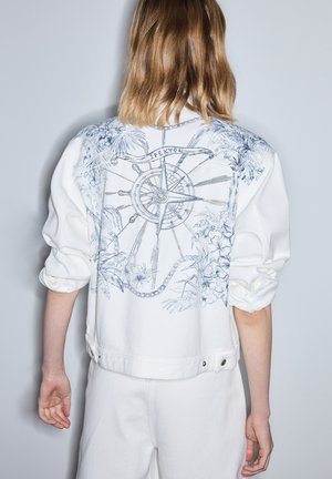 Osoba o włosach sięgających do ramion, ubrana w białą kurtkę z niebieskim motywem marynistycznym kompasu i kwiatowym na plecach, w połączeniu z białymi spodniami.