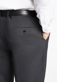 Pantaloni grigi scuri con una texture liscia, dotati di una tasca posteriore con chiusura a bottone e una cintura in pelle nera. La camicia è bianca.