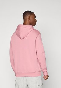 Hoodie en coton rose avec une capuche à cordon, des poignets et un ourlet côtelés, arborant une étiquette sur le côté. Porté avec un short cargo gris.